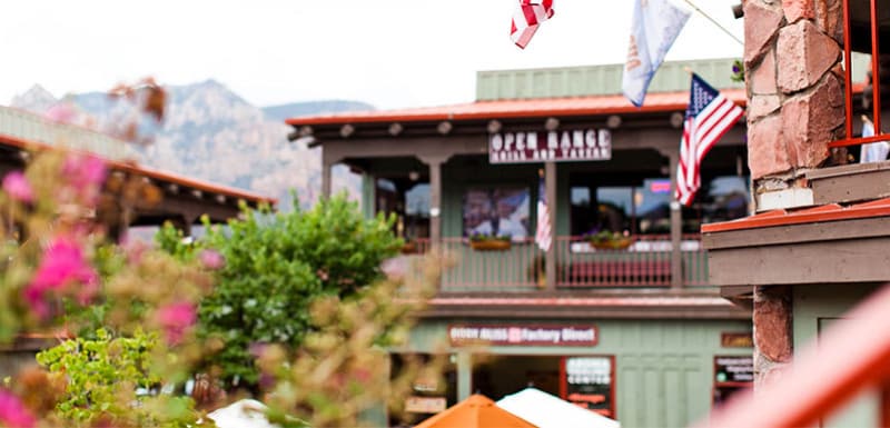 Живописные фасады зданий центра города Седоны с цветущими цветами на переднем плане, зданием с вывеской «OPEN RANGE GRILL AND TAVERN» и величественными горами на заднем плане.